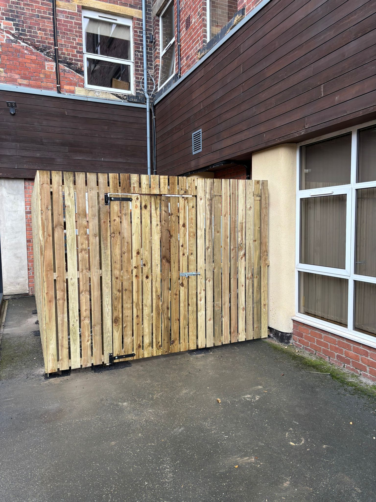 Wooden gate blocking a narrow outdoor passage beside a brick building and window.