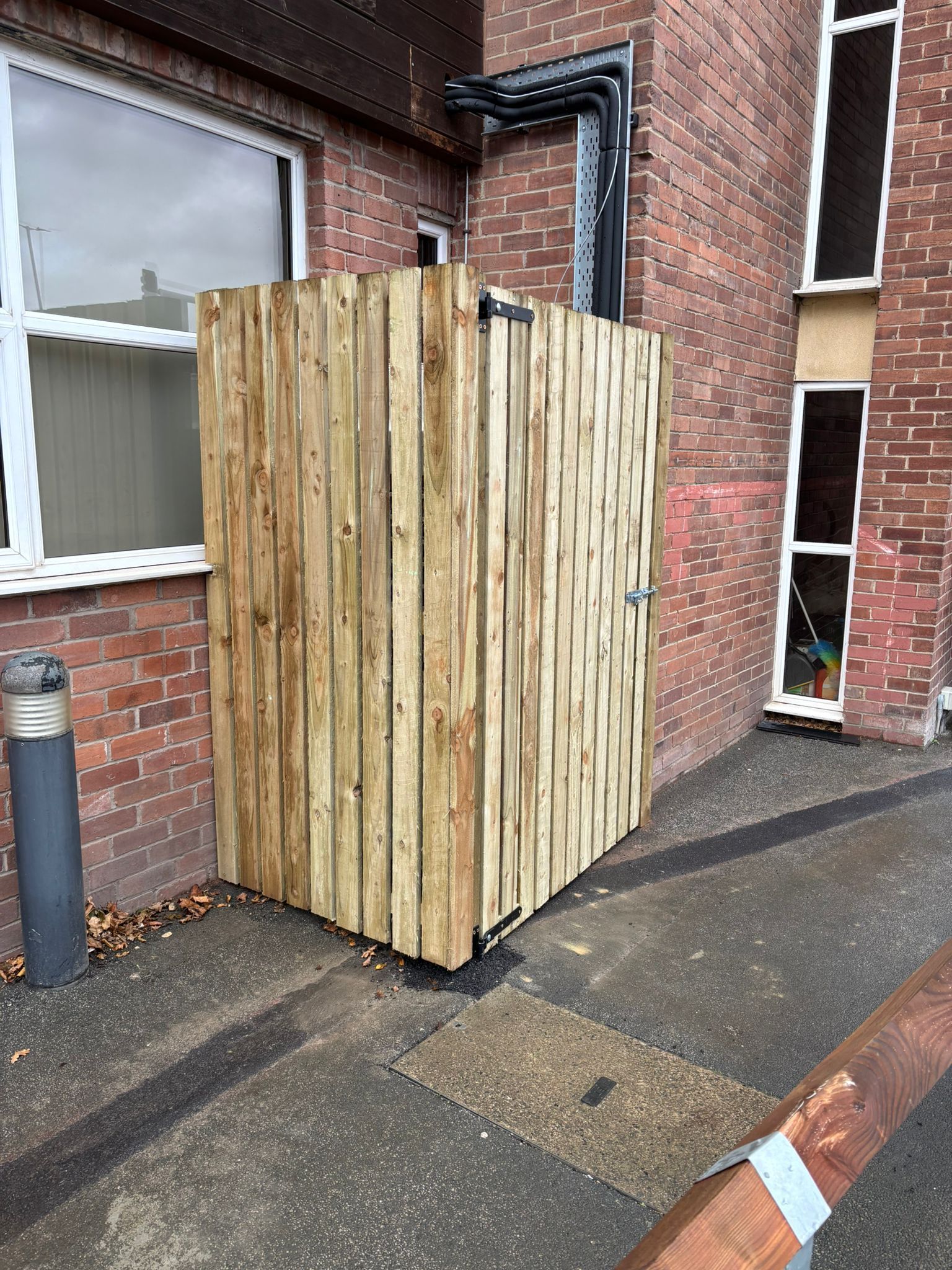 Tall wooden enclosure beside a brick building on a wet sidewalk.