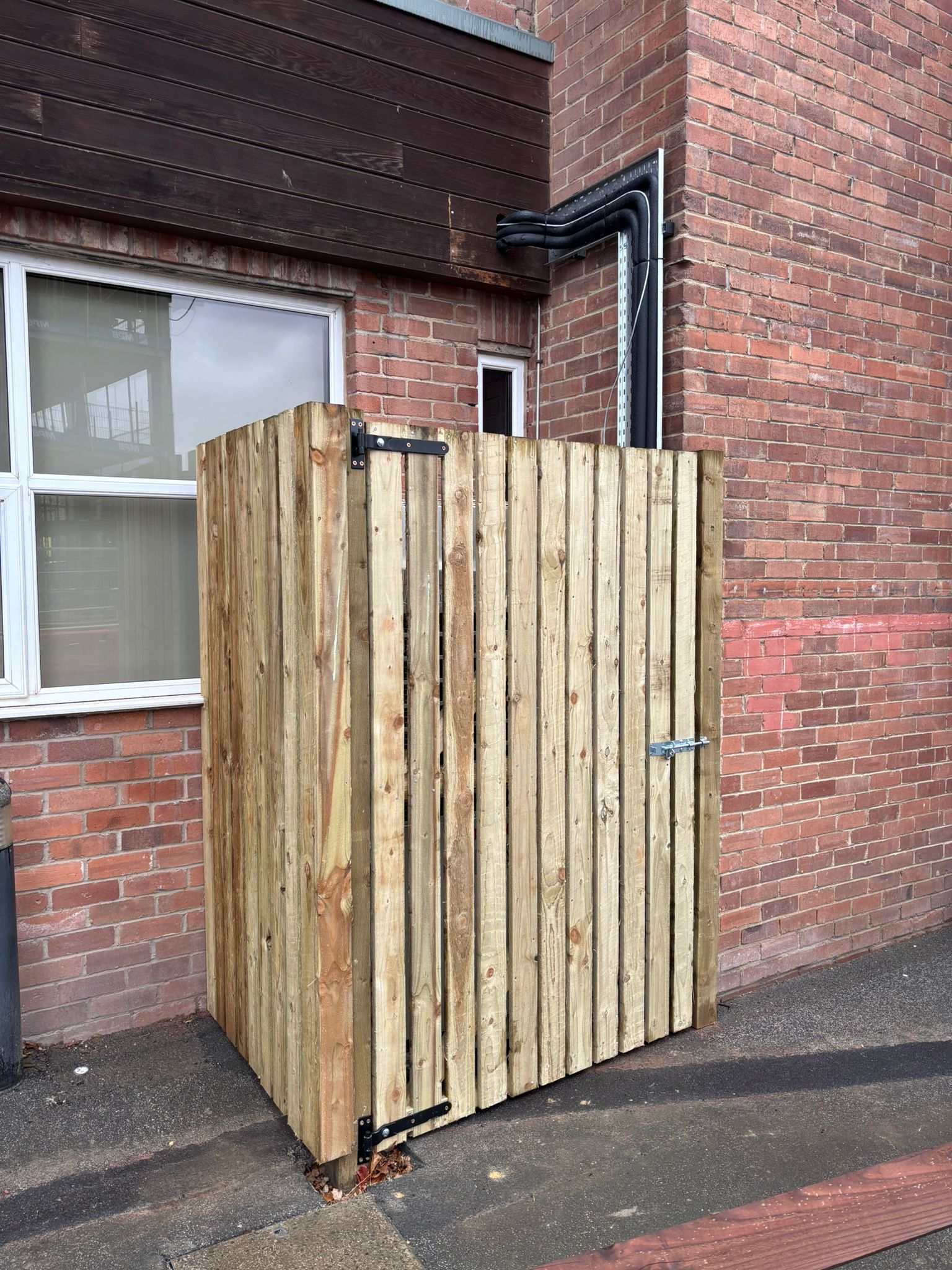 Wooden enclosed structure beside a brick building, with a window and drainpipe nearby.