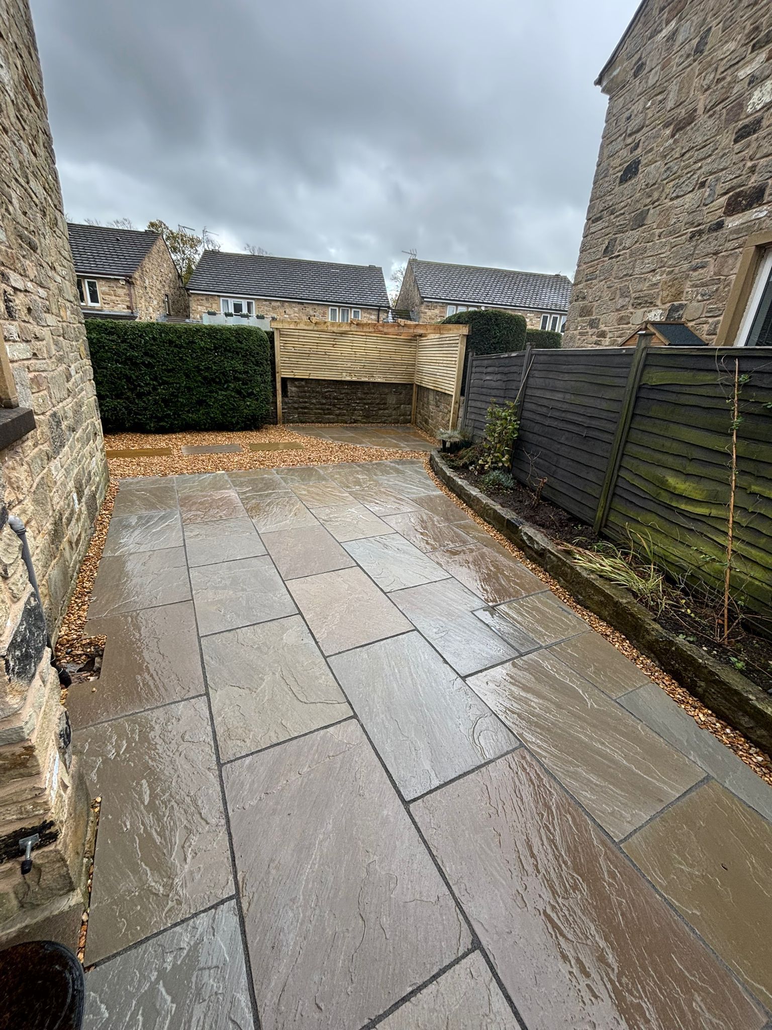 Wet stone patio between stone buildings, with fence and hedge under a cloudy sky.