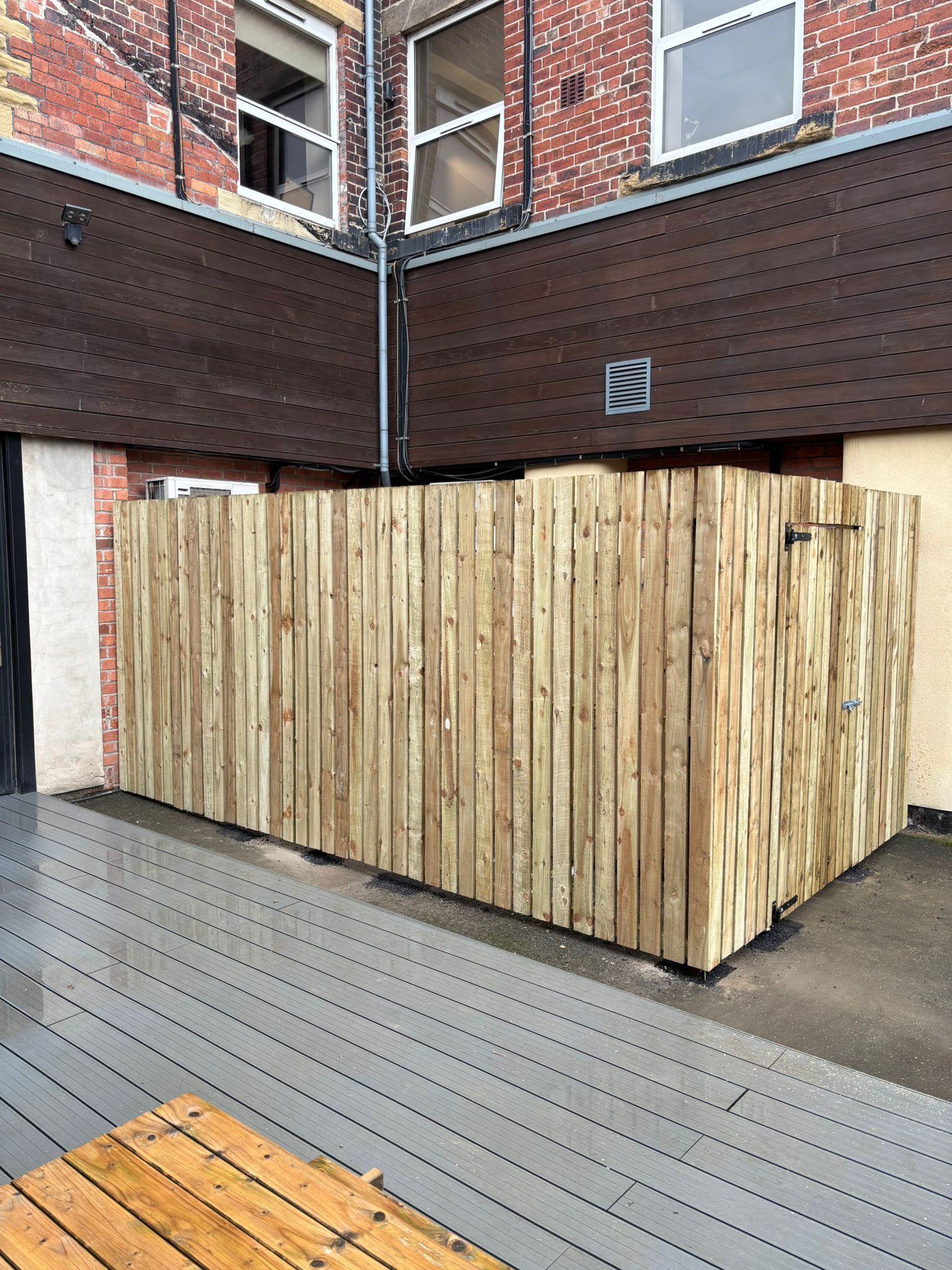 Wooden dumpster enclosure beside a brick building on a gray paved courtyard