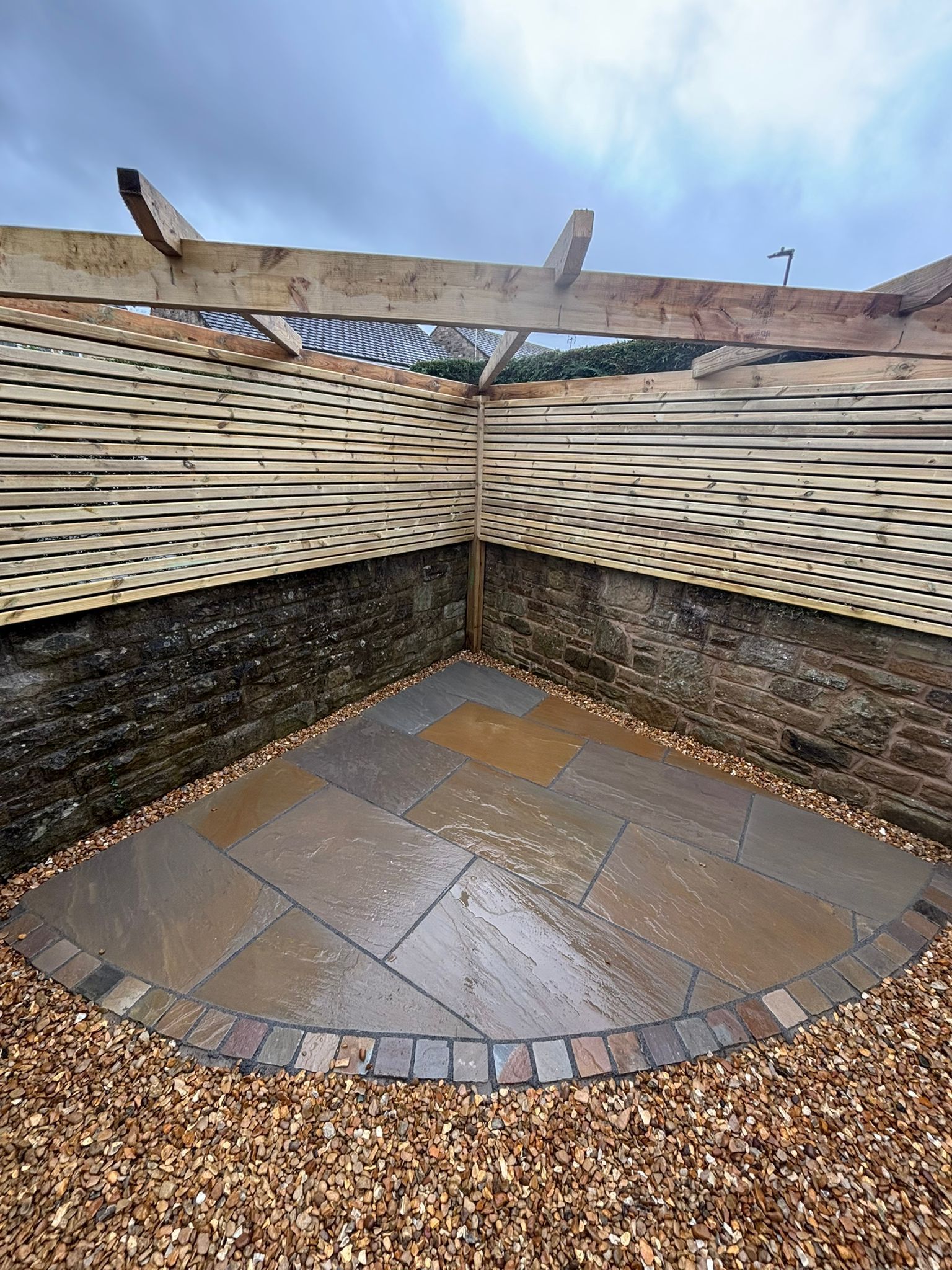 Small paved corner seating area with woven wood walls, gravel border, and a rustic beam overhead