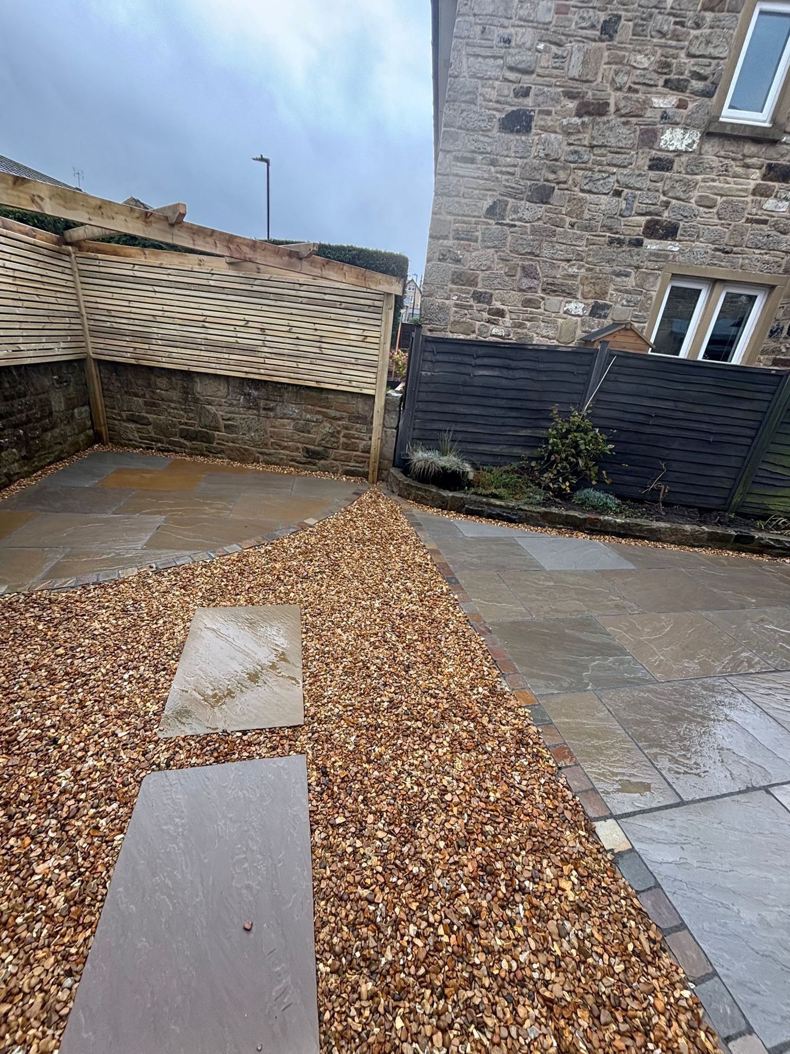 Stone courtyard with wet pavement, scattered leaves, and a stone building with a wooden fence and blue sky.