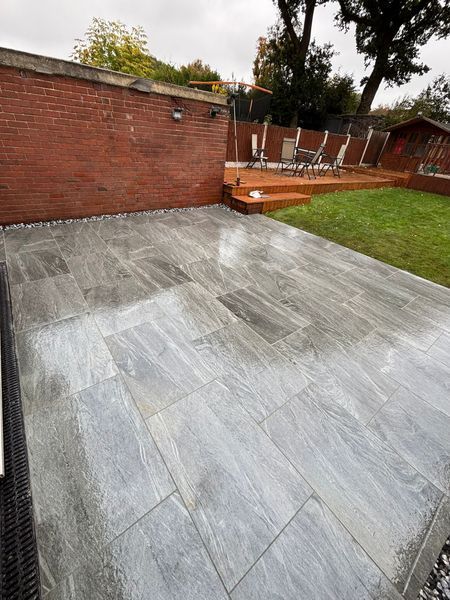 Backyard patio with gray stone paving, red brick wall, lawn, and wooden raised seating area with chairs