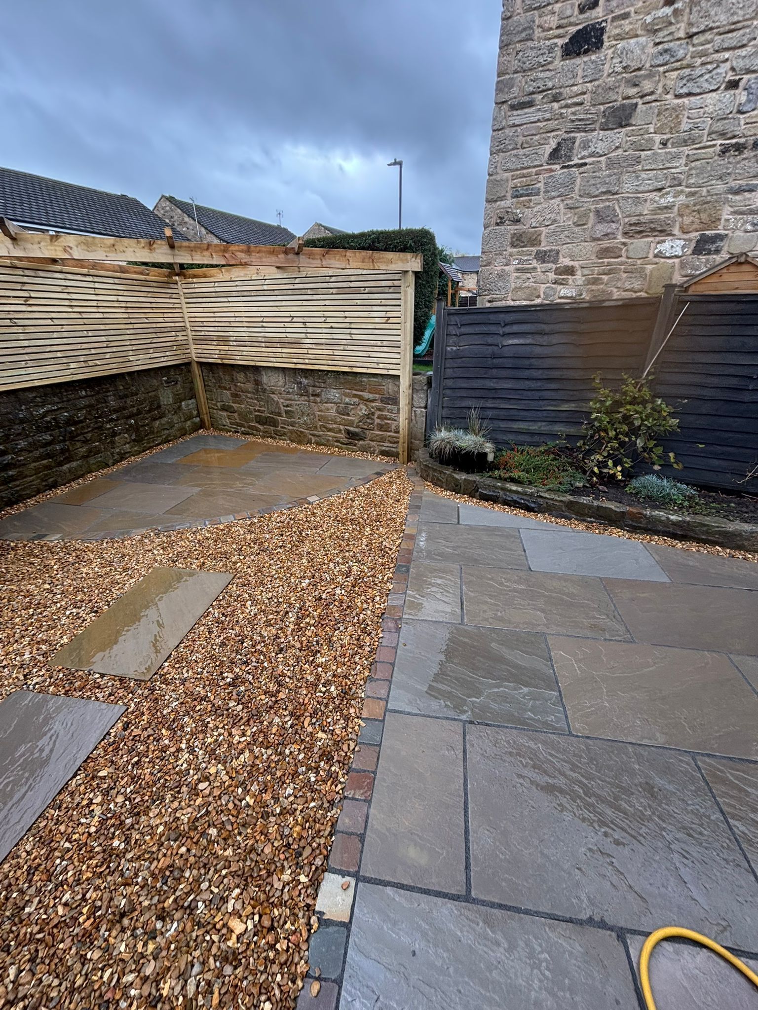 Small paved courtyard with gravel path, wooden fencing, stone wall, and hose on the ground