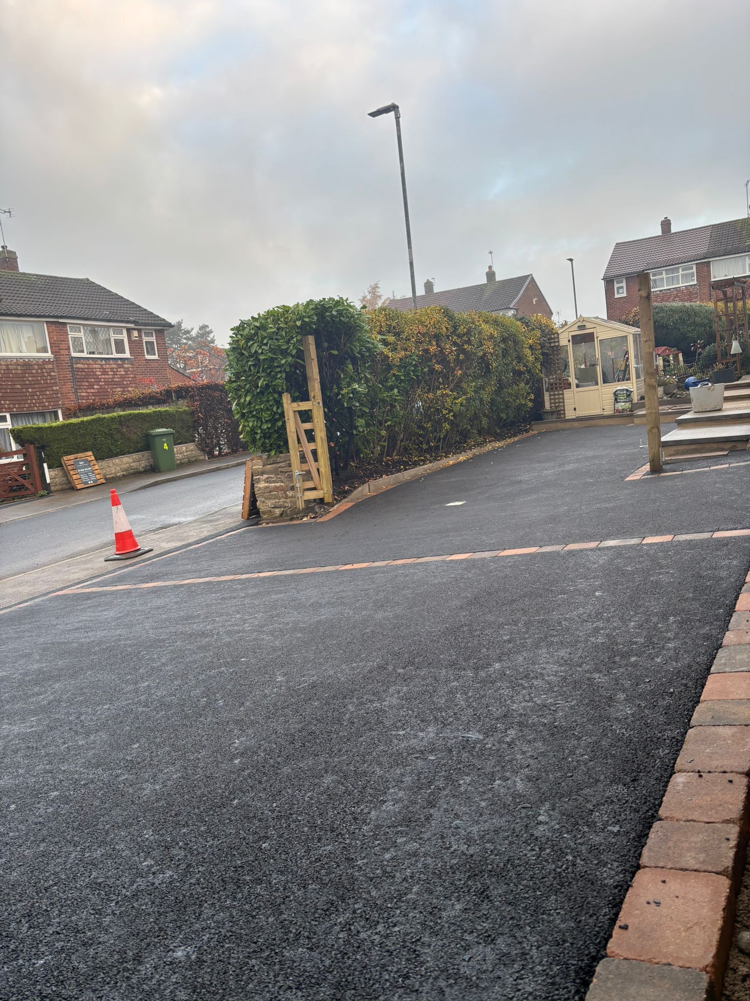 Quiet residential street with a freshly paved driveway, hedge, parked car, and orange cone near a gate.