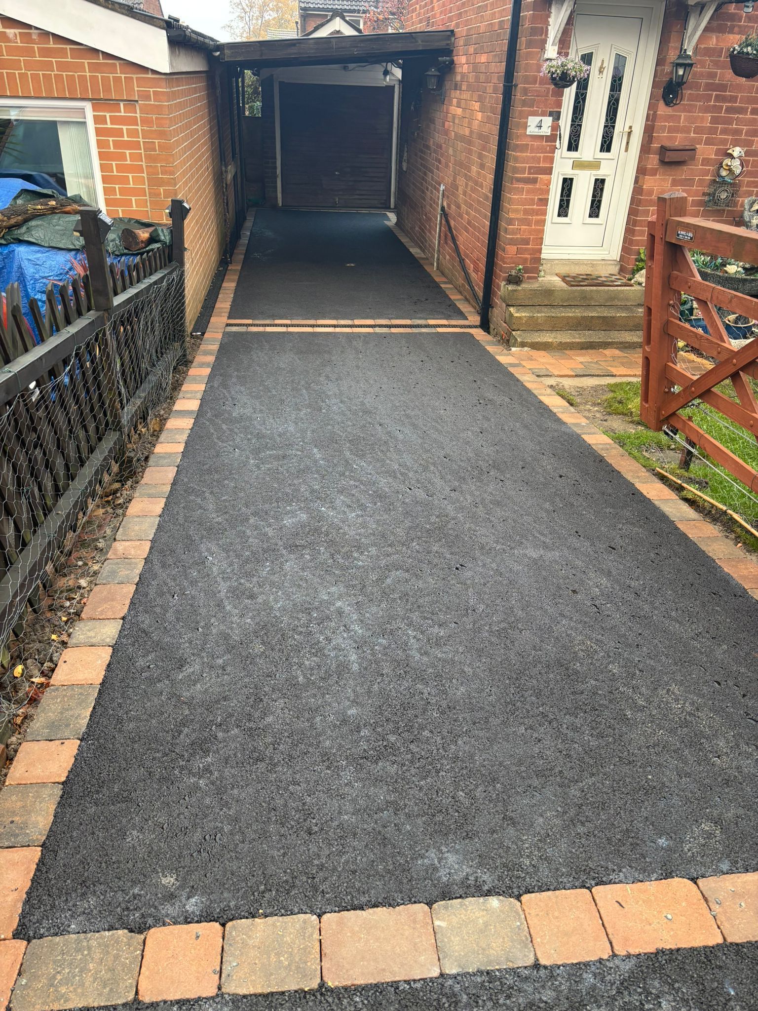 Newly paved black driveway leading to a garage, bordered by brick and wooden fences.