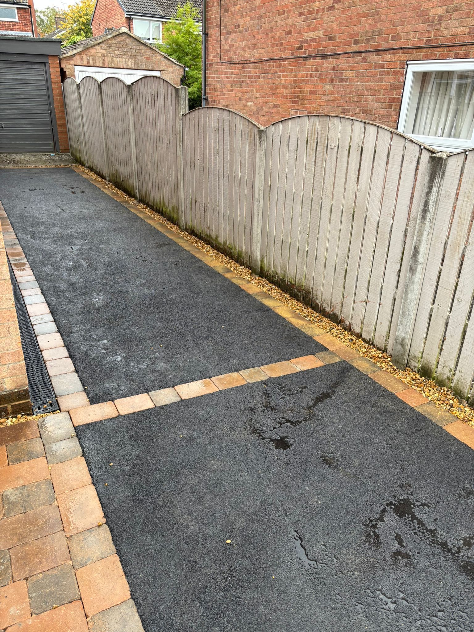 Narrow paved driveway beside a wooden fence and brick walls, with a small shed at the end.