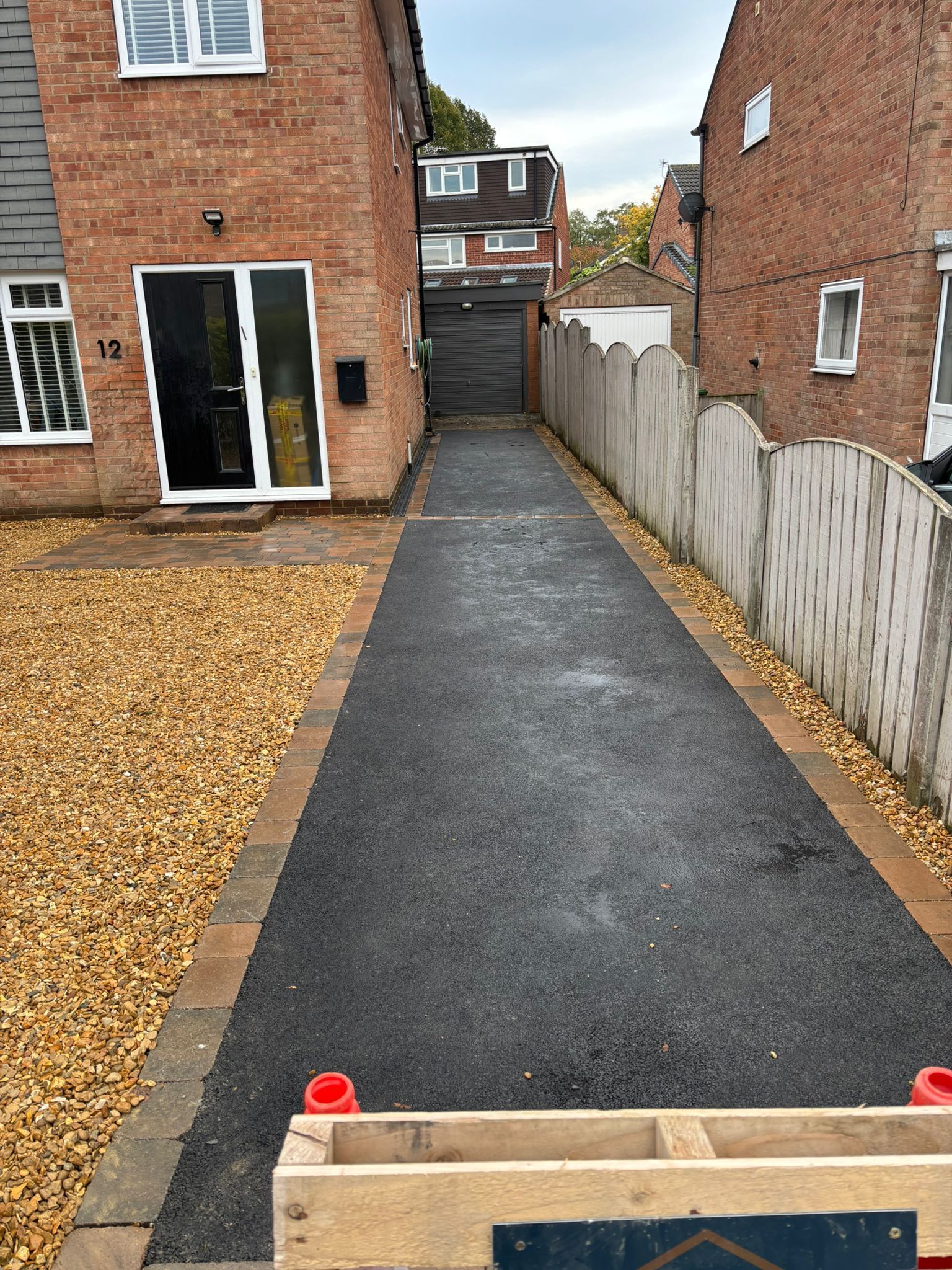 Fresh black asphalt driveway between brick houses, bordered by gravel and a white fence.