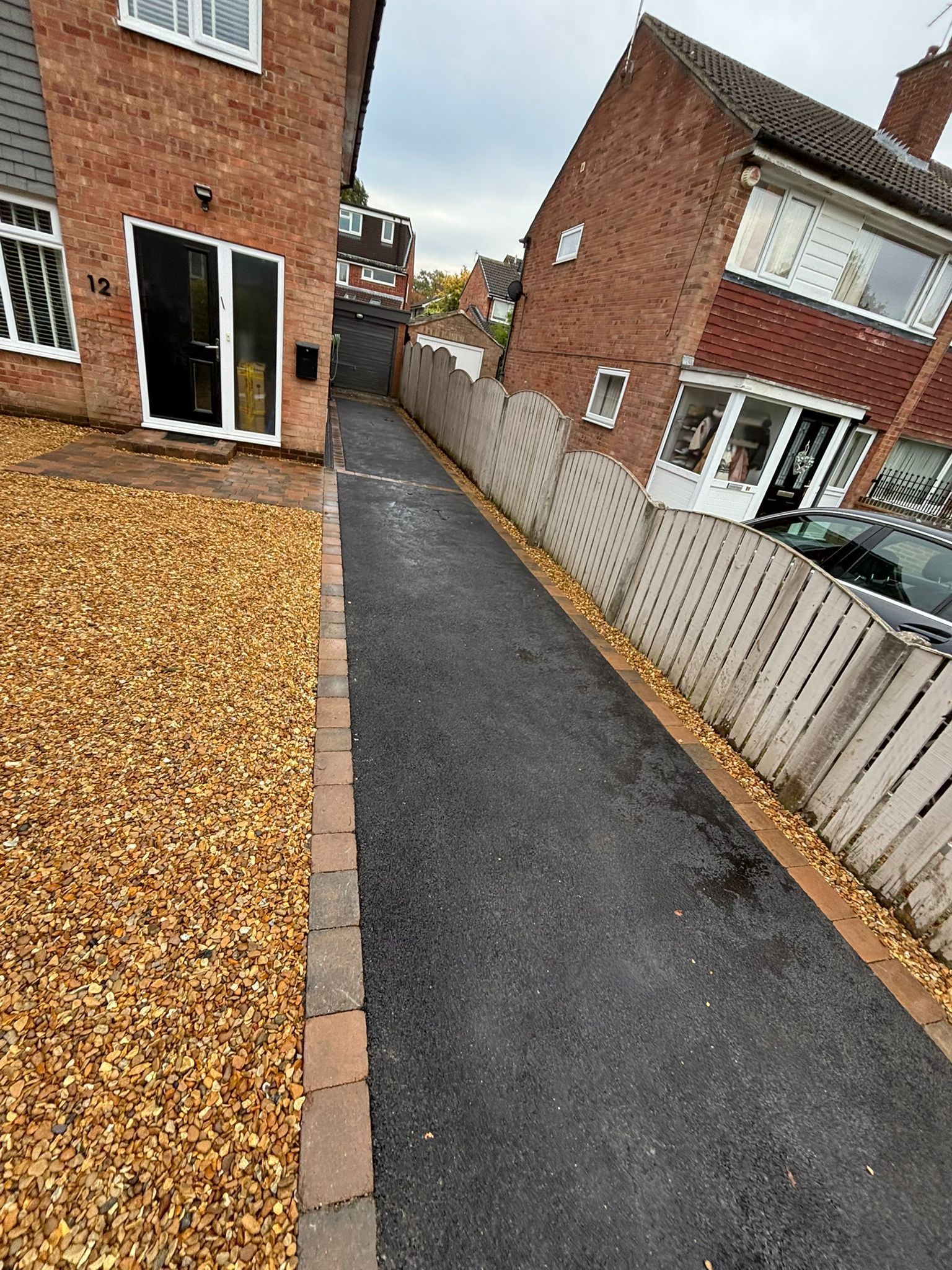 Narrow paved side path between brick houses, with gravel on one side and a fence on the other.