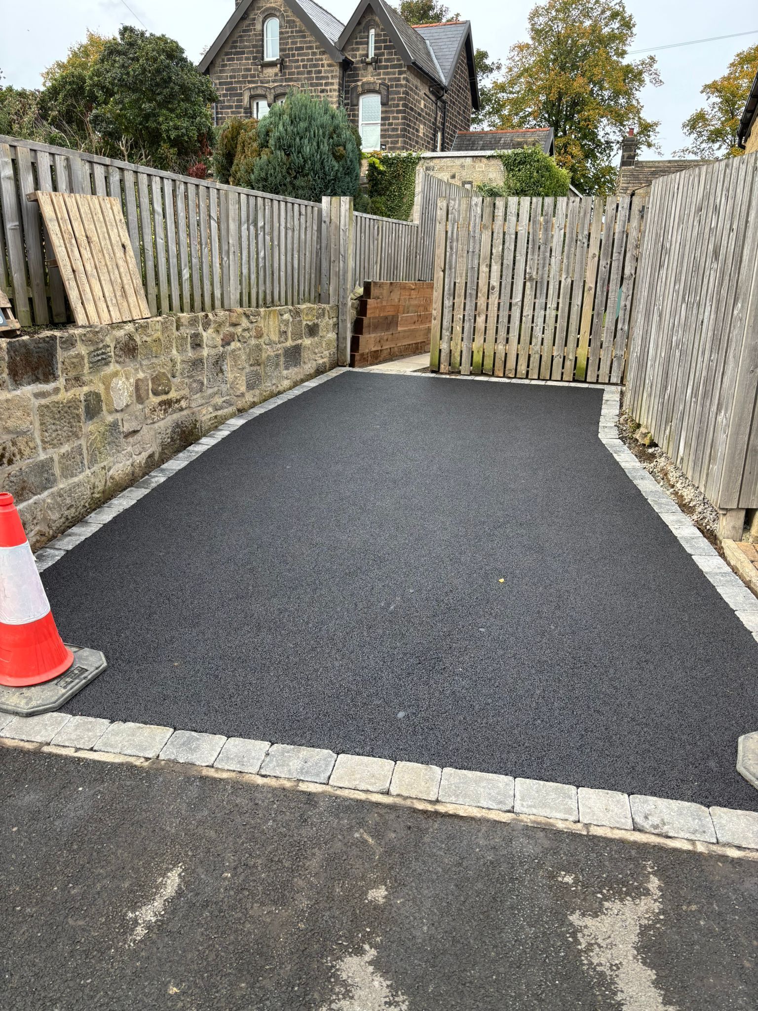Freshly paved asphalt driveway beside stone walls, wooden fence, and a traffic cone at the entrance