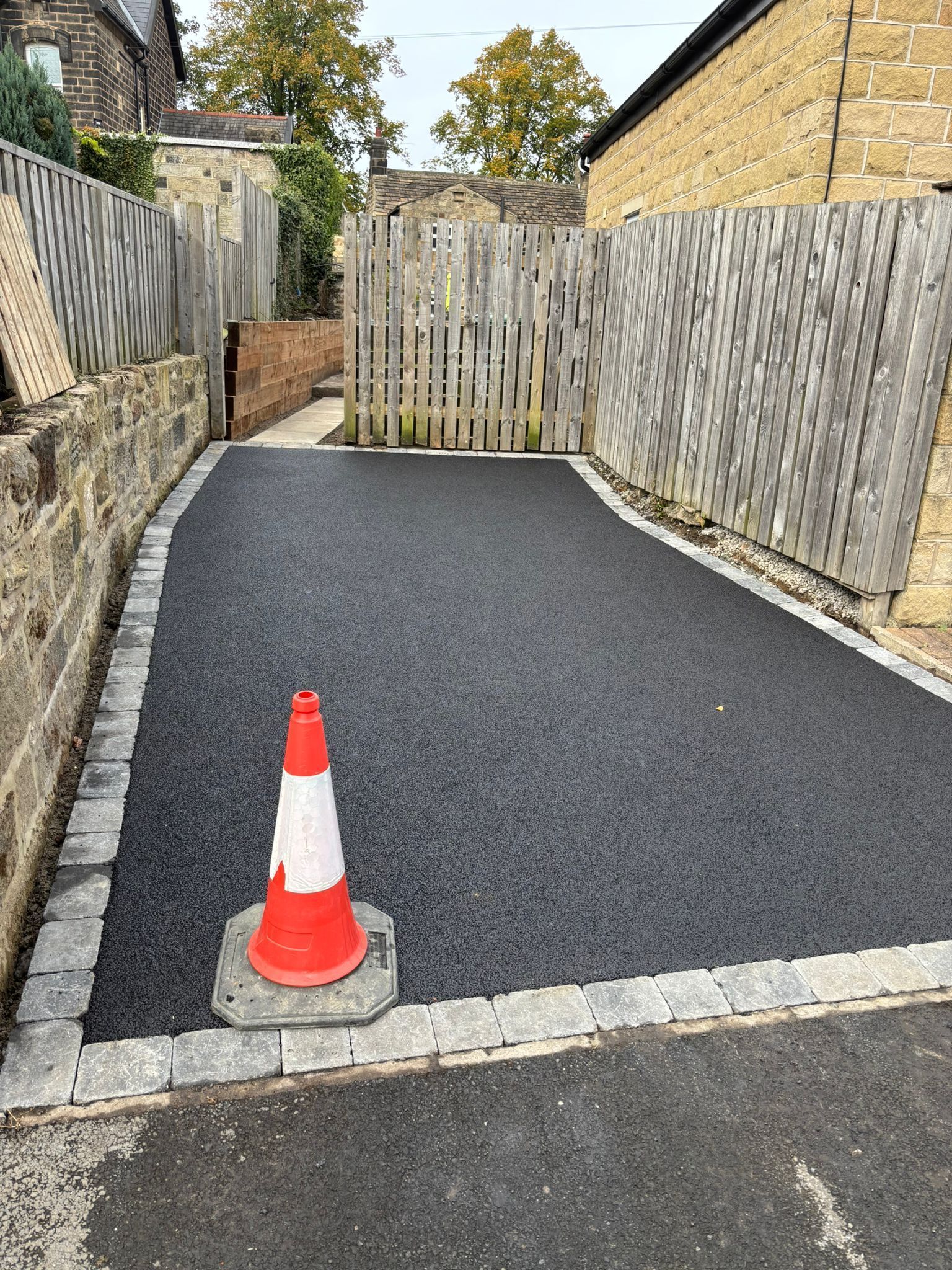 Freshly resurfaced narrow driveway or path with black asphalt, white edging, and an orange traffic cone in front