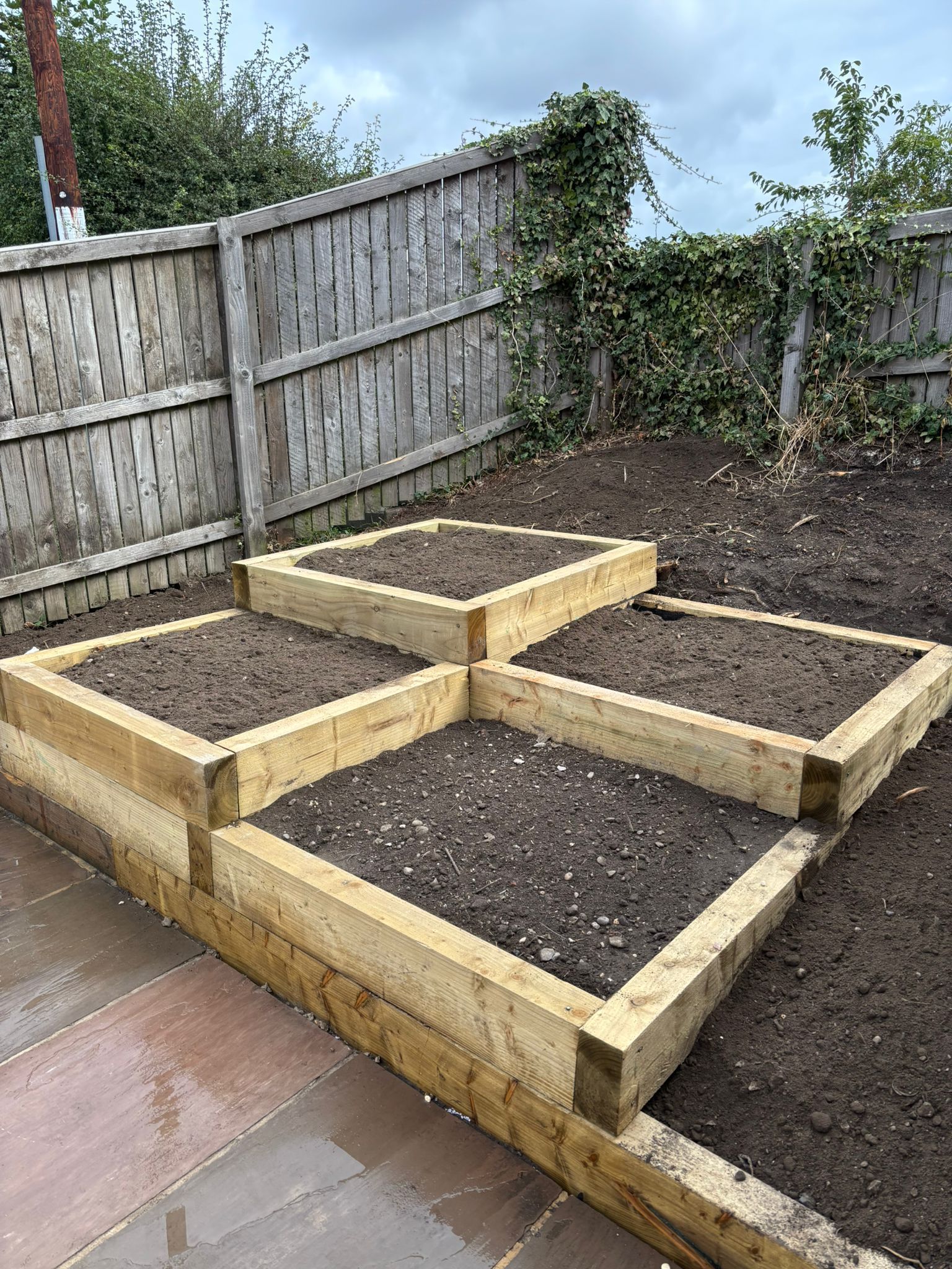 Four raised wooden garden beds filled with soil in a backyard beside a fence and patio.