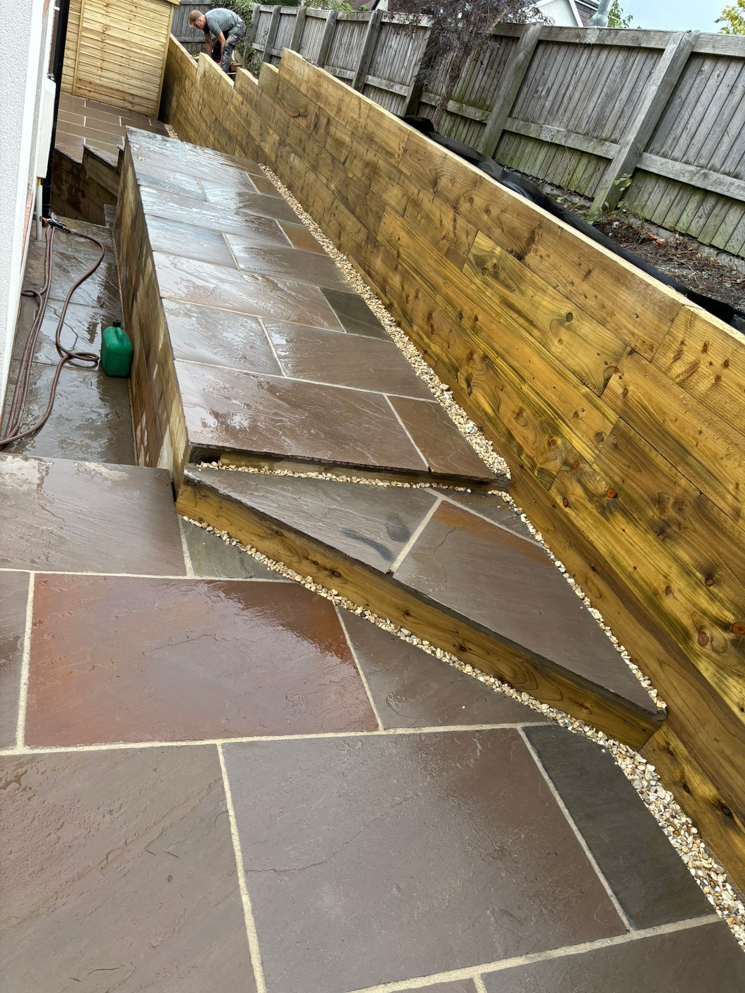 Damaged sloped walkway with cracked concrete slabs beside a wooden fence after rain.
