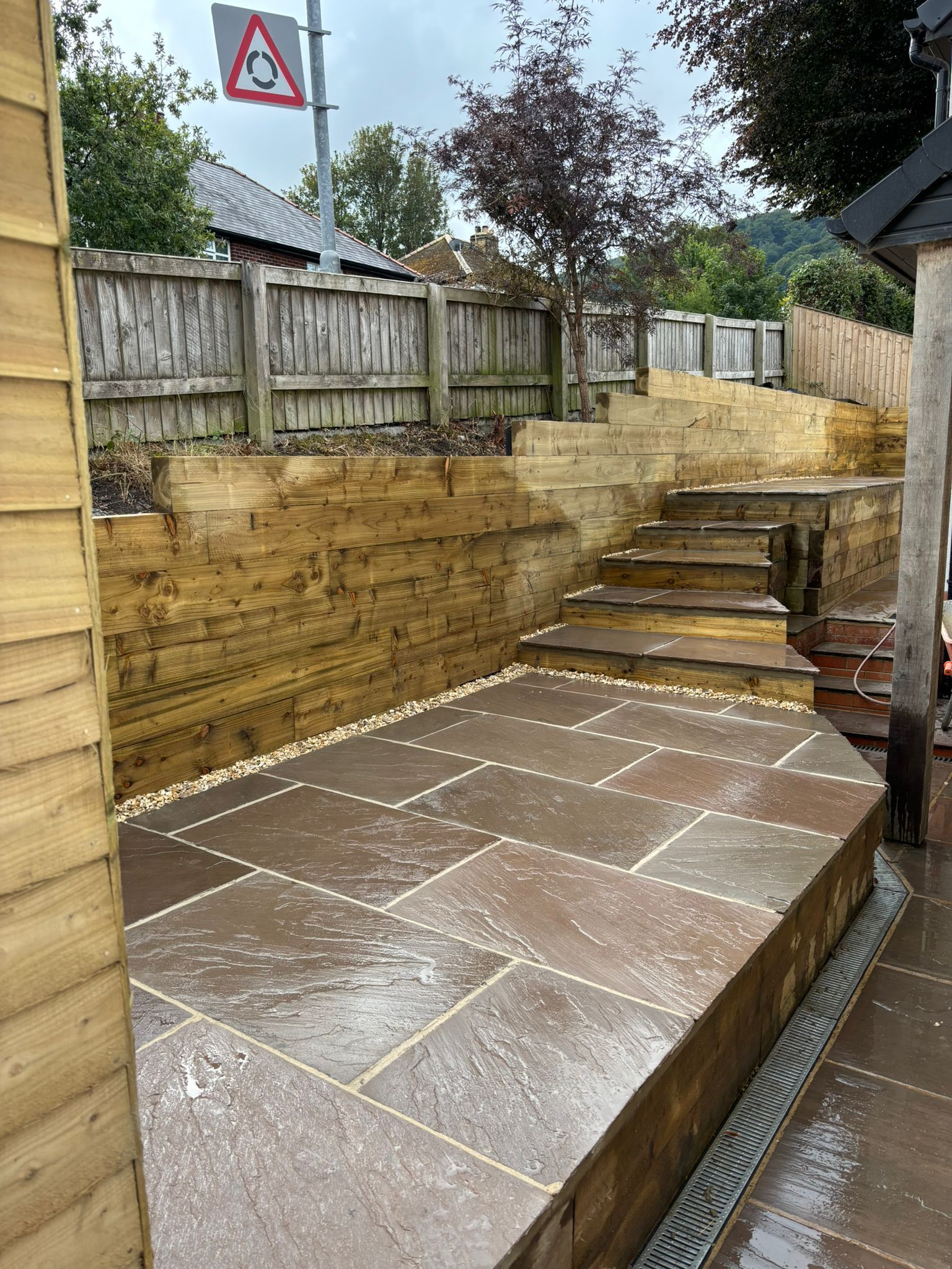 Wet stone patio beside tiered steps and retaining walls in a fenced backyard.