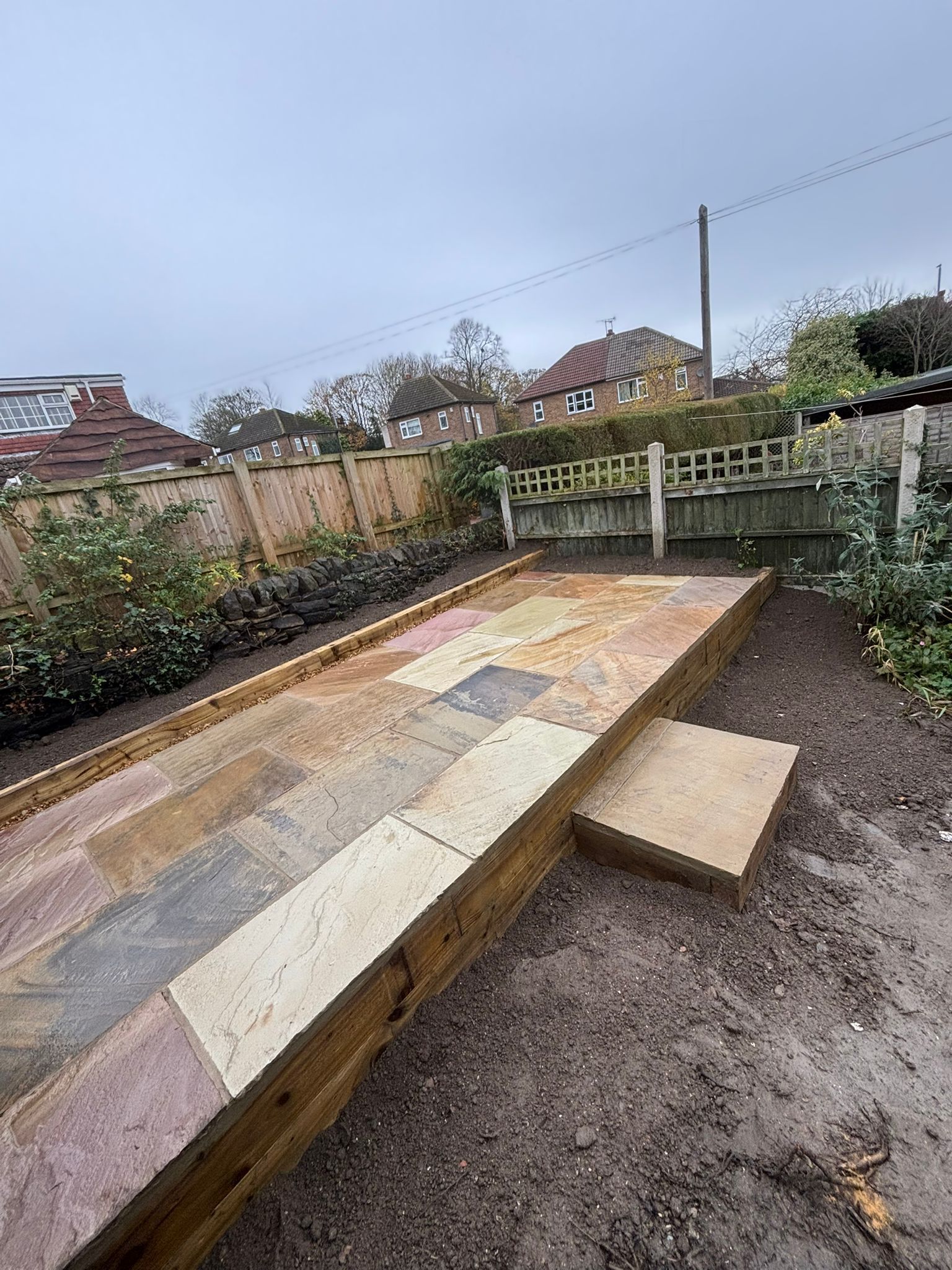 Backyard with a wooden raised platform and patchwork paving in a muddy garden.
