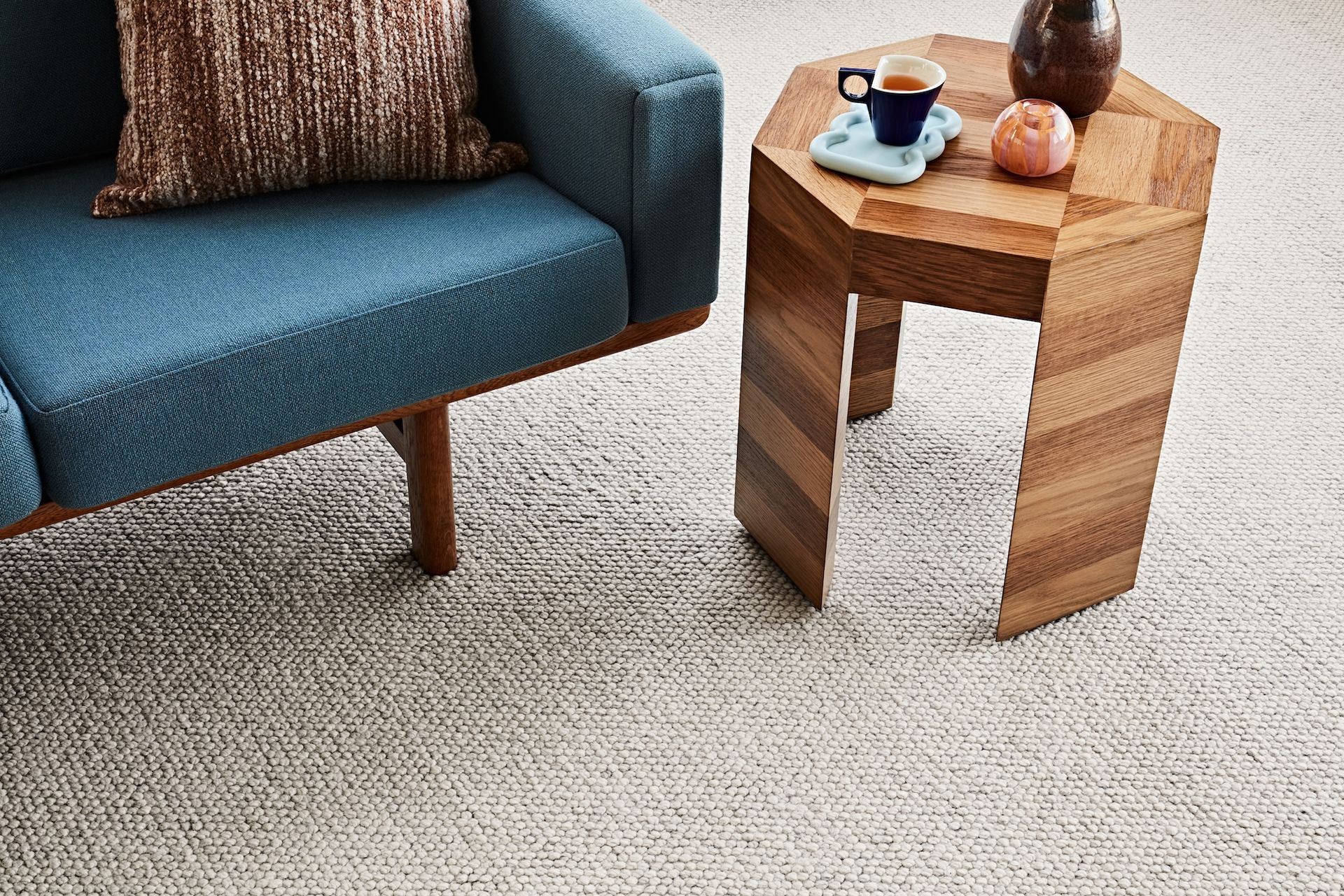 Teal couch with brown pillow, a geometric wood side table with a cup, and a light rug.