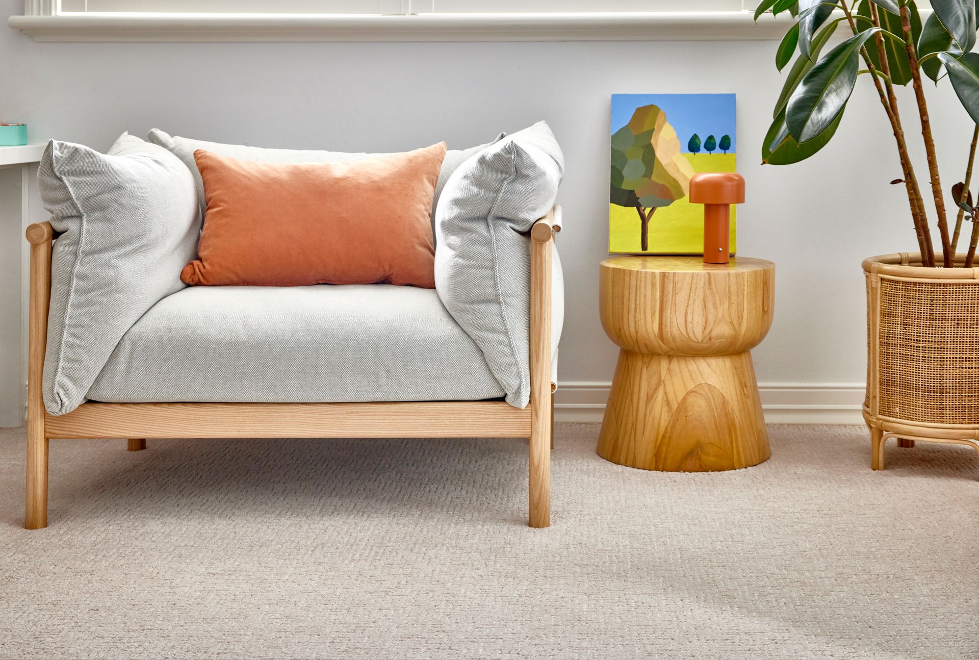 Cozy living room with a white armchair, orange pillow, wooden side table, painting, and potted plant on light carpet.