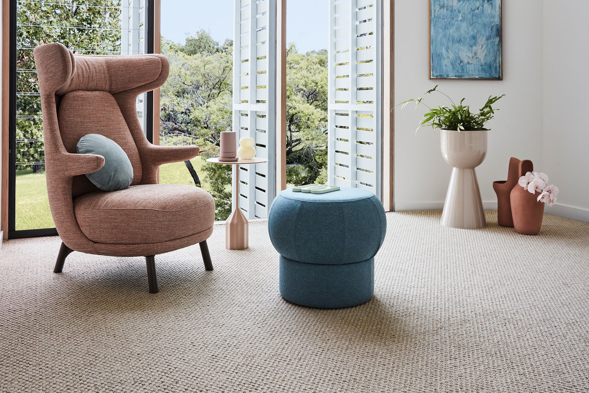 A cozy living room with a pink armchair, blue ottoman, and patterned carpet. Large windows overlook lush greenery.