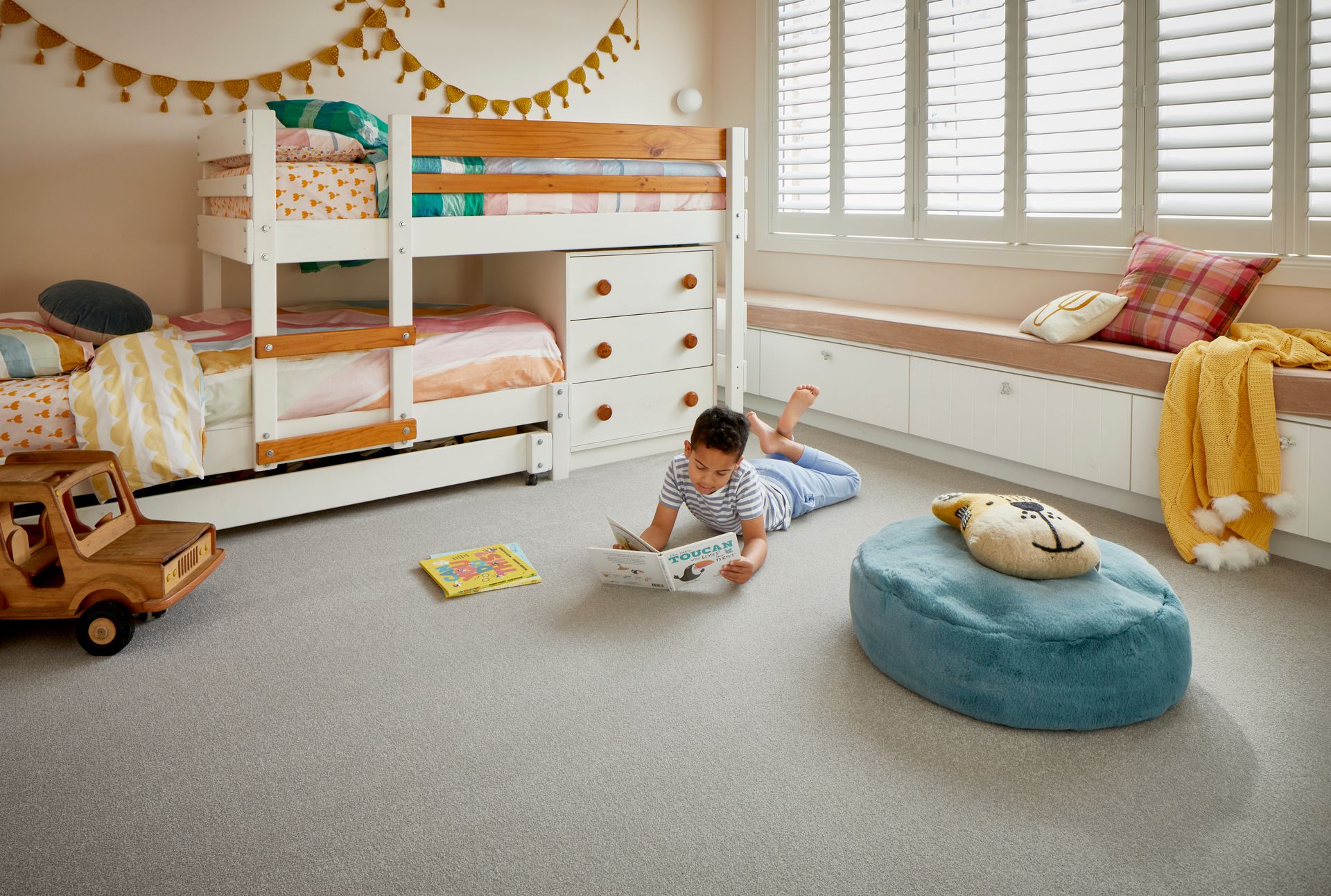 A child reads on a gray carpeted floor in a bedroom. A bunk bed, window seat, and plush ottoman are also present.
