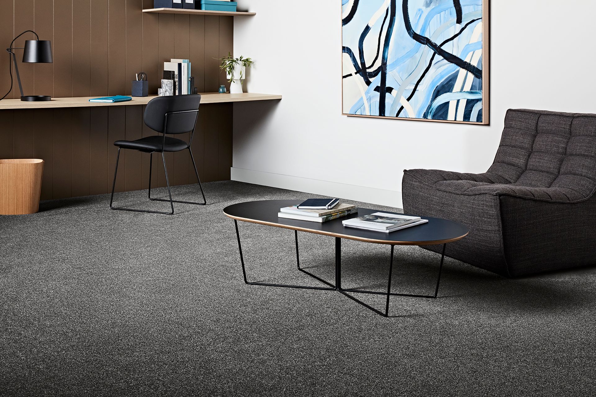 A modern office space with a desk, chair, and abstract painting, featuring a low coffee table, and a textured armchair over speckled carpet.