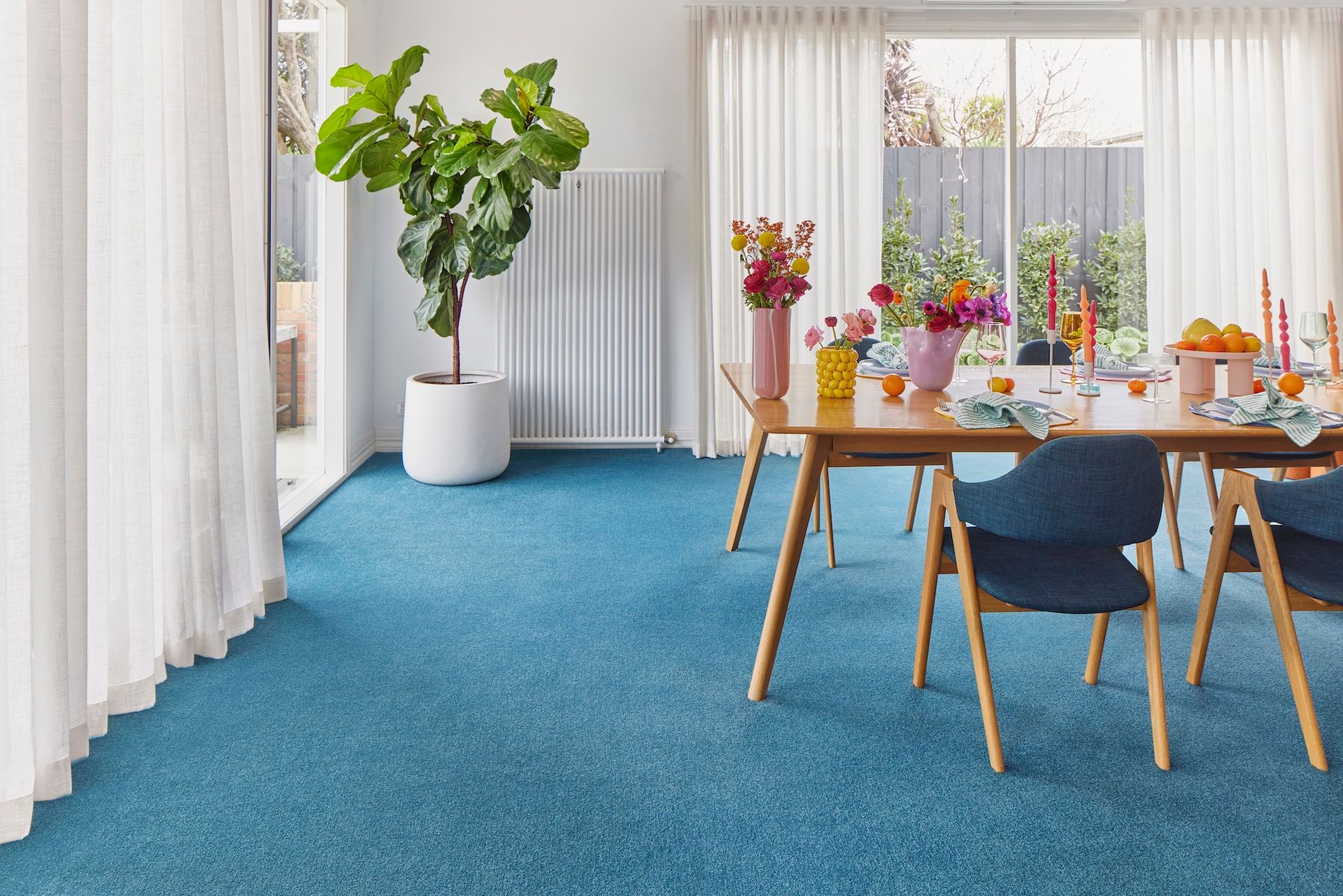 A dining room with a blue carpet, wooden table, colourful flowers, and large plant. Soft curtains frame a window with an outdoor view.