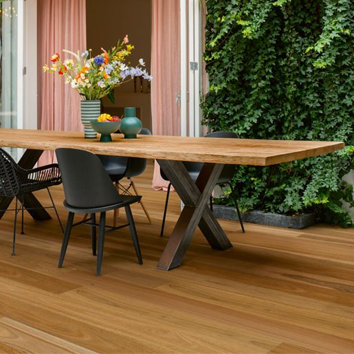 Wooden outdoor dining table with black chairs on a wood deck, flowers and climbing vines in the background.