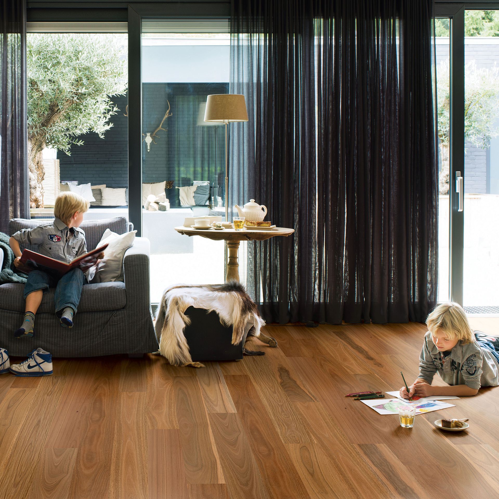Two young boys in a living room with hardwood floors. One reads on a couch, the other draws on the floor; dark curtains and sliding glass door.