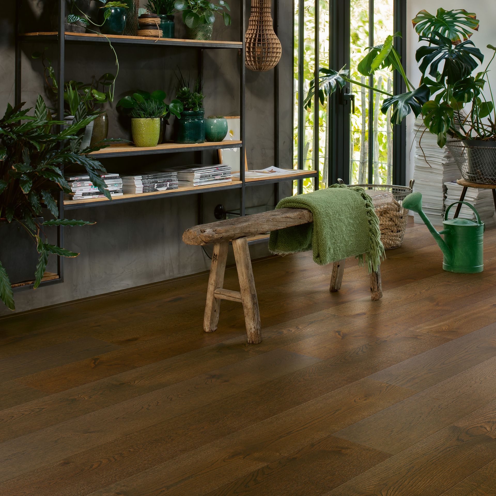 Wooden floor in a room with plants. A wooden bench has a green blanket, and shelving with plants.