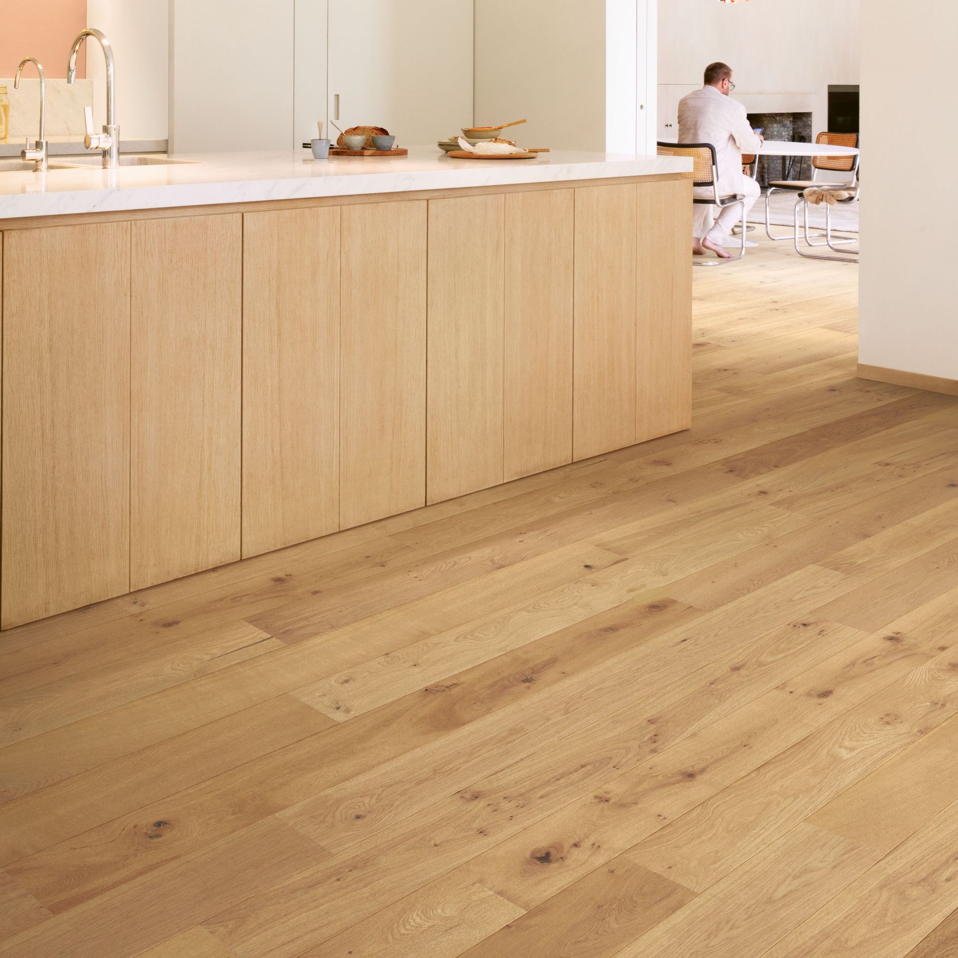 Wooden floor with a kitchen island and a person in the background. The island and cabinets are light wood.