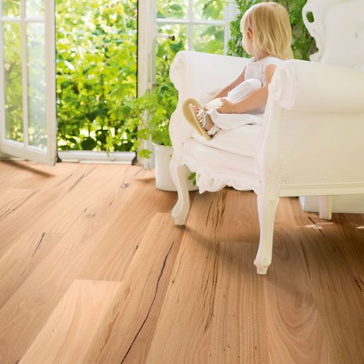A young child sits in a white armchair near an open window overlooking greenery, on a light-coloured wood floor.