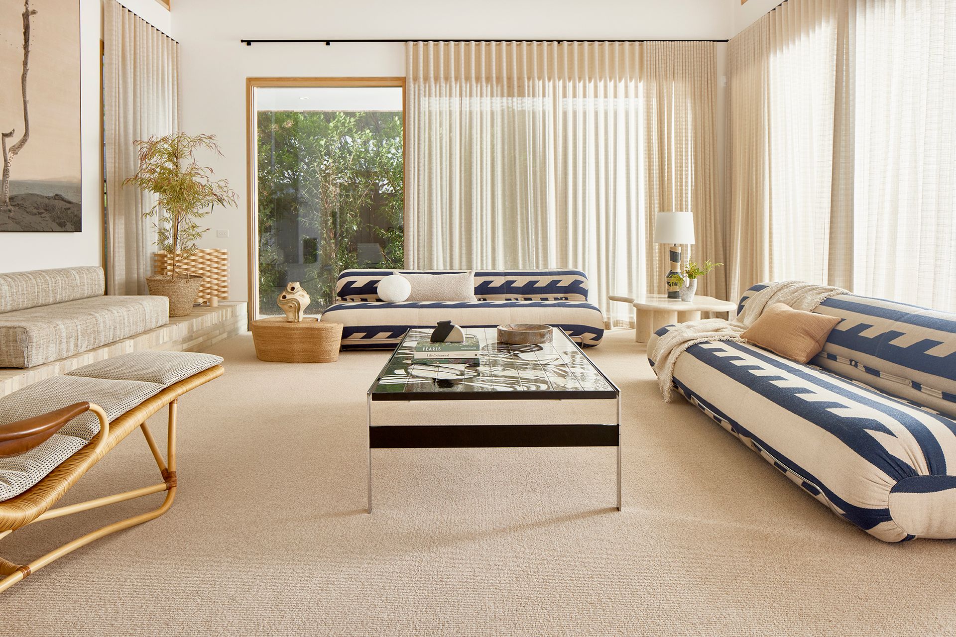 A bright living room with two patterned sofas, a glass coffee table, and large windows with sheer curtains.