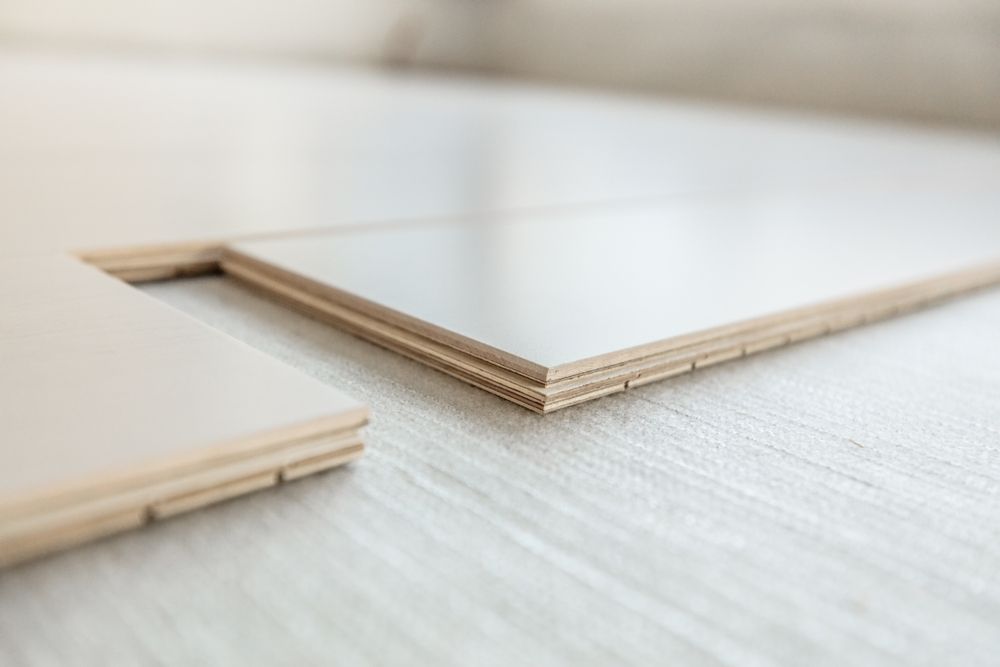 Close-up of light-coloured laminate flooring planks with interlocking edges, lying on a textured white surface.