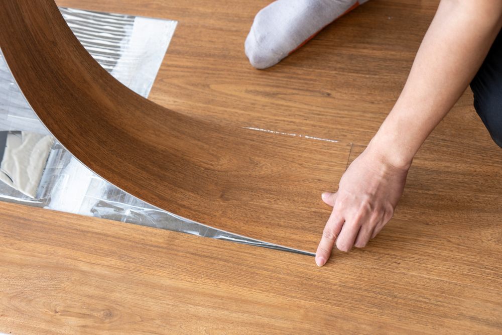 Person installing vinyl plank flooring, pointing finger at the floor edge. One foot with a sock is visible.