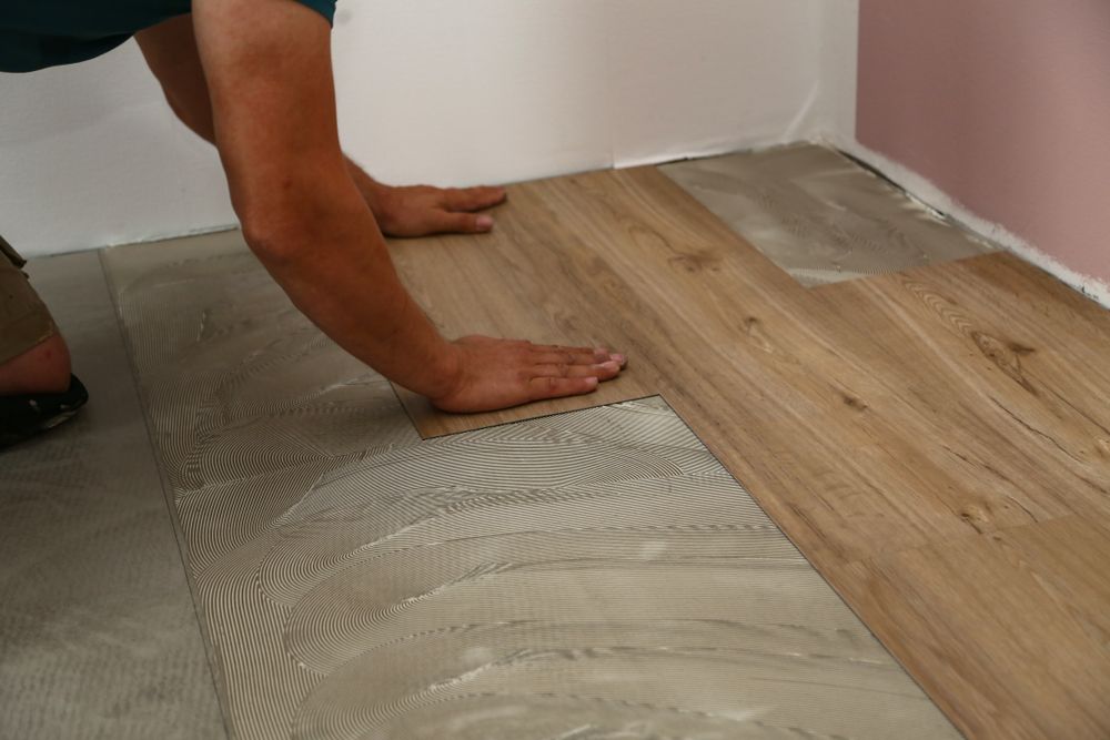 A person installing wood-look flooring near a wall, placing the planks. The floor is light brown; the walls are white and pink.