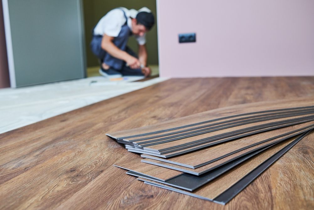 A worker installing wooden laminate flooring in a room with a pink wall; a stack of flooring boards is in the foreground.