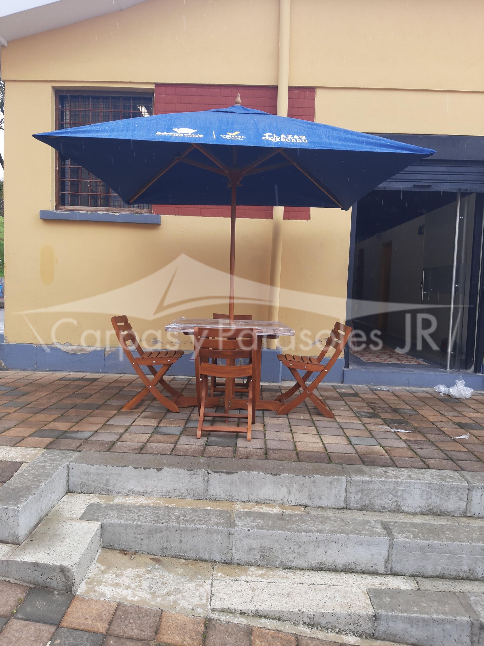 Una sombrilla cuadrada azul cubre una mesa de madera y tres sillas plegables en un patio pavimentado frente a un edificio.