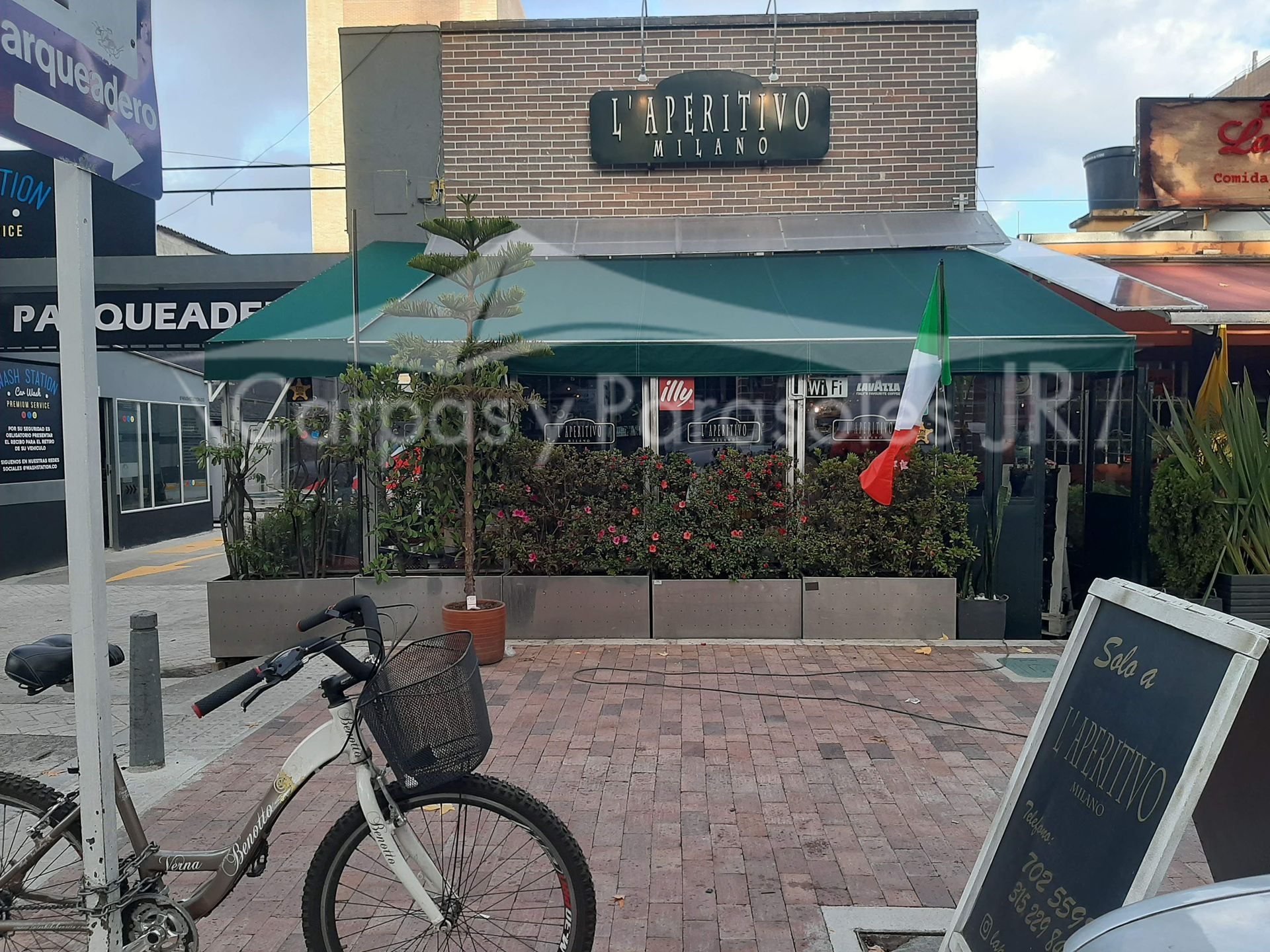 Una bicicleta está parada frente a una cafetería con toldos verdes, banderas italianas y un cartel en la acera.