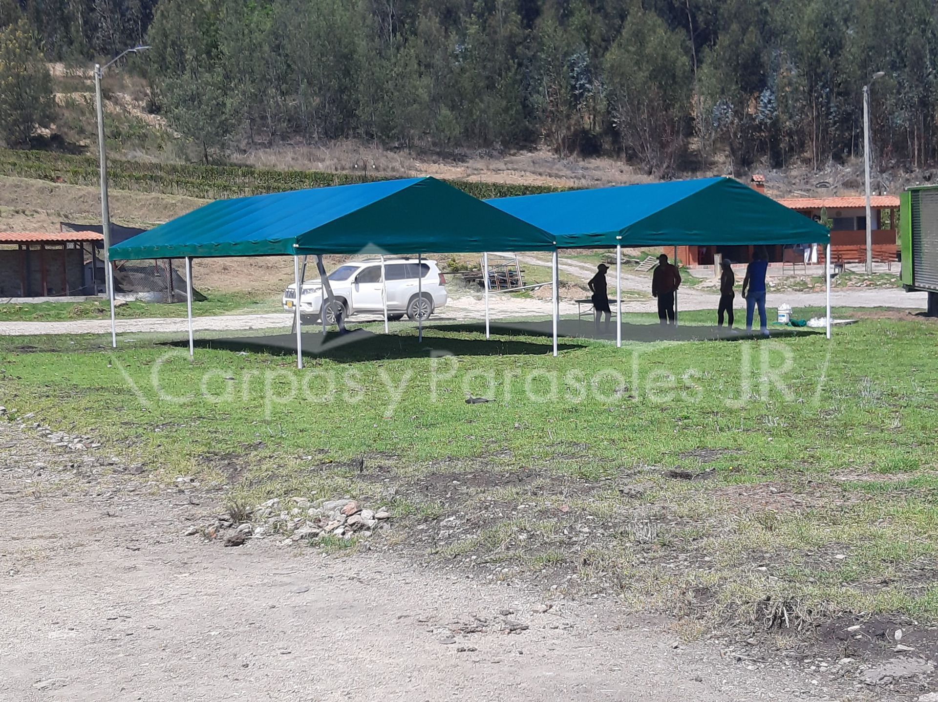 Dos grandes carpas de exterior de color verde azulado instaladas en un campo de hierba, con un SUV blanco y gente cerca.