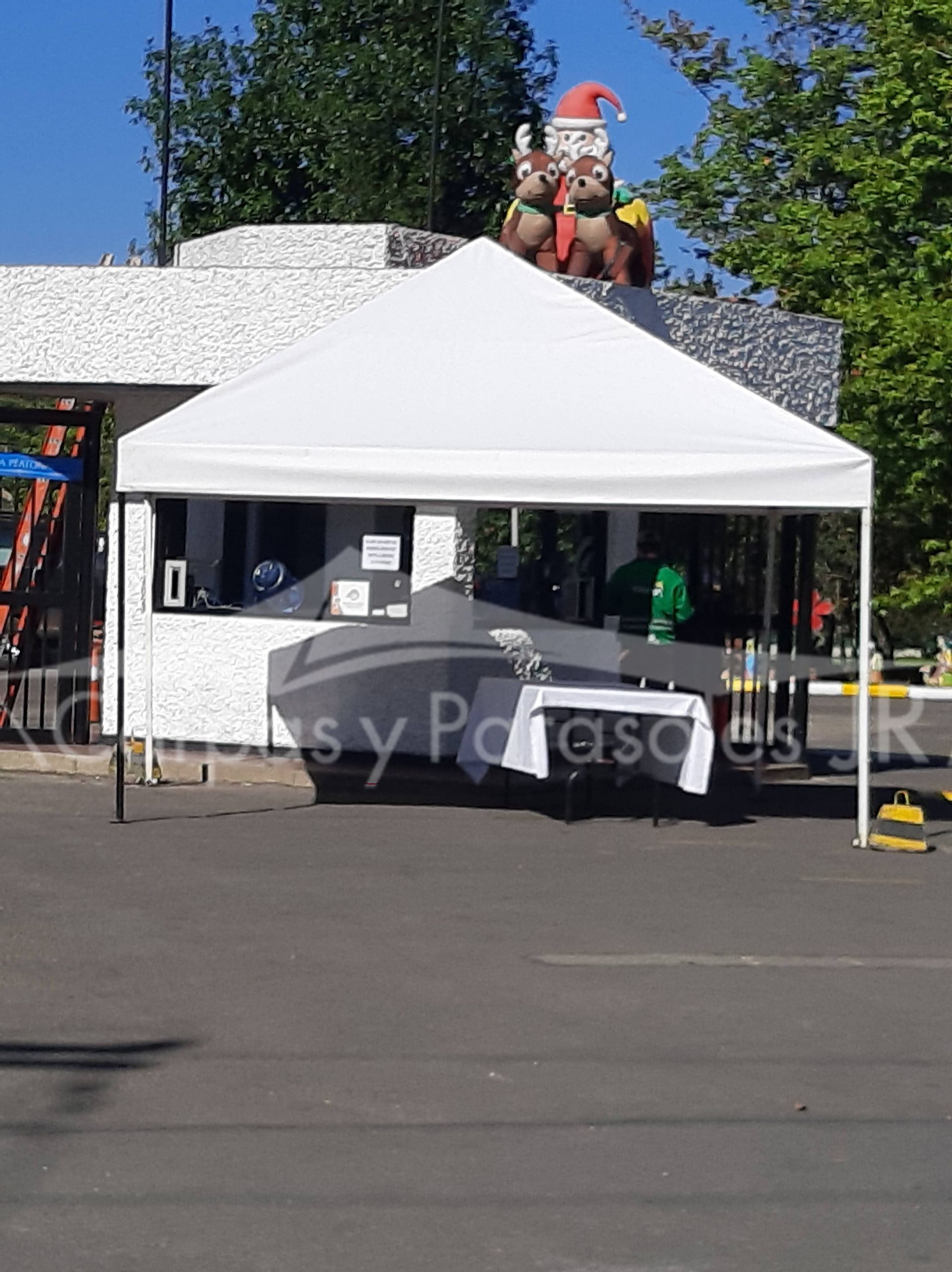 En un aparcamiento hay una carpa blanca plegable, con una mesa debajo y una decoración de Papá Noel en el edificio de detrás.