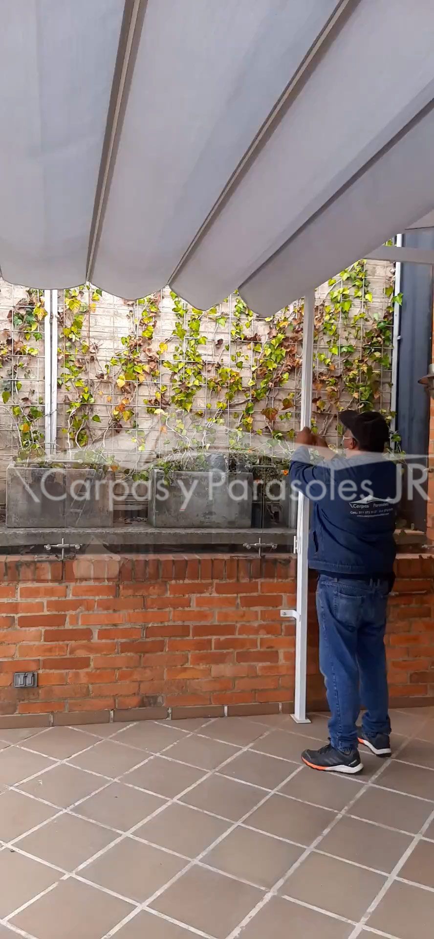 Un trabajador vestido de azul instala un toldo retráctil blanco sobre un patio con una pared de ladrillo
