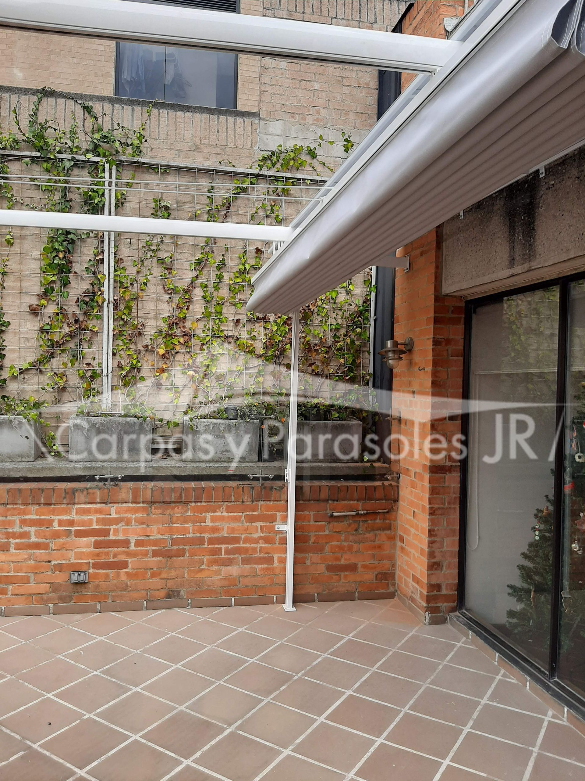 Un toldo retráctil blanco se extendía sobre una terraza embaldosada con una pared de ladrillo y plantas trepadoras verdes.