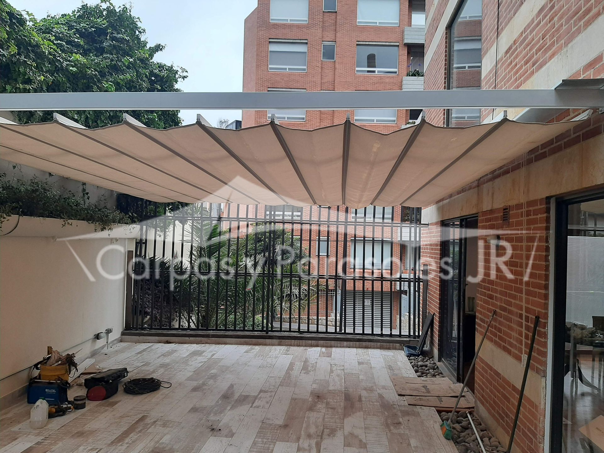 Una pérgola retráctil de color beige instalada sobre un balcón con baldosas, junto a un edificio de ladrillo rojo.
