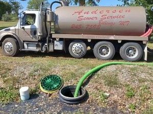 A vacuum truck from Anderson Sewer Service parked on grass with a green hose inserted into an open septic tank riser.