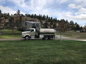 A water delivery truck is parked on a driveway in a rural setting, with a hose extending across a green lawn.