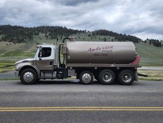 A brown and white Andersen Excavating tank truck driving on a road against a backdrop of hills and a cloudy sky.