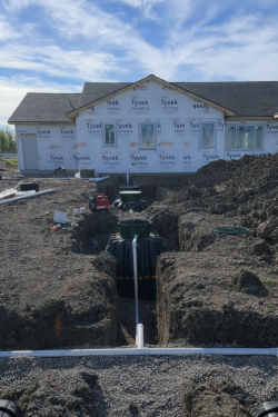 A construction site with a new house wrapped in white Tyvek, featuring a buried septic tank and trench pipes in the ground.