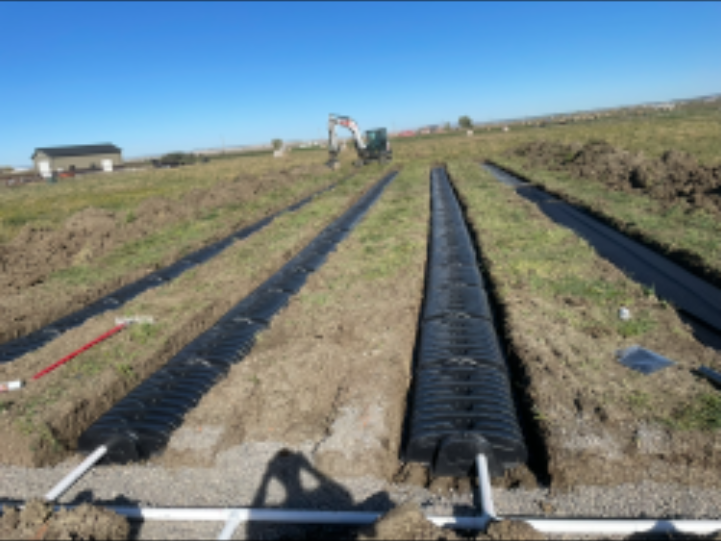 An excavator installs rows of black plastic septic drain field chambers in a field with white PVC piping.