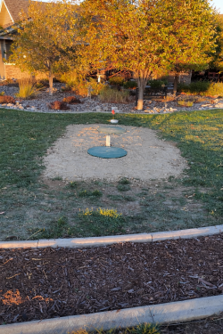 A green septic tank lid with a white pipe sits centered in a patch of gravel surrounded by grass in a backyard.