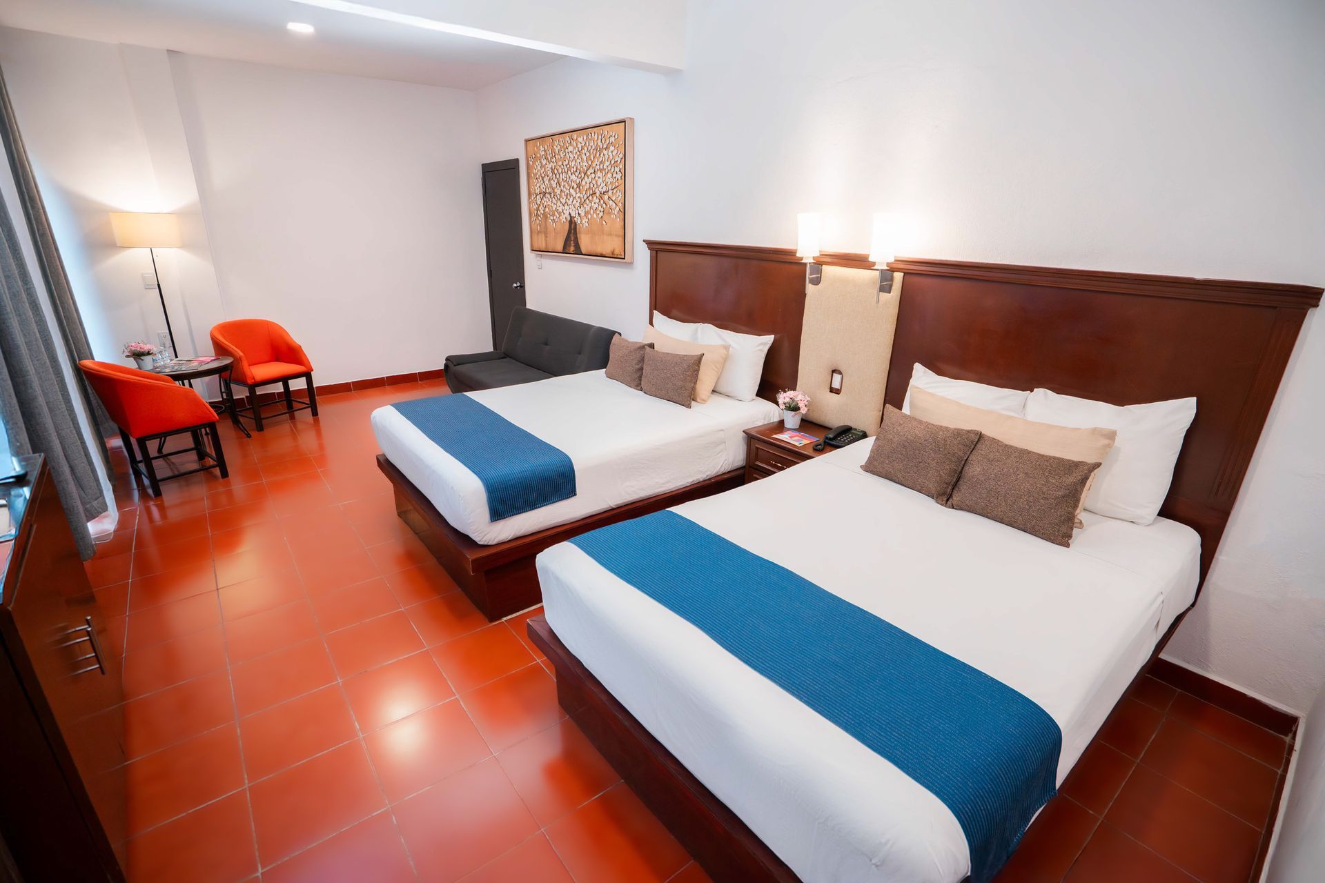 Habitación de hotel con dos camas dobles, ropa de cama azul y blanca, sillas naranjas y un pequeño sofá.